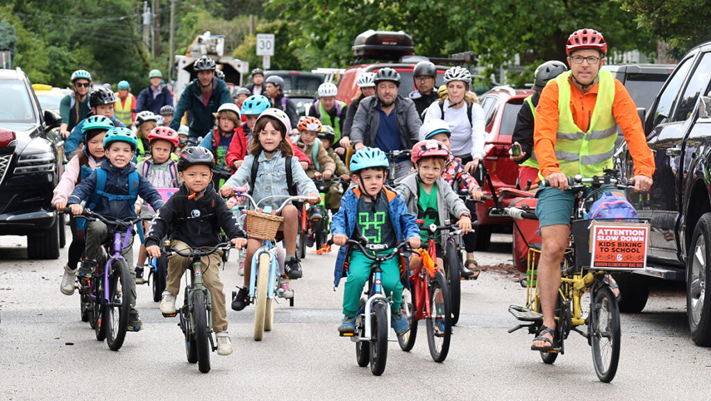 A large group of school-aged children riding bicycles together down a street in a Bike Bus, accompanied by a few adults who are also on bicycles.