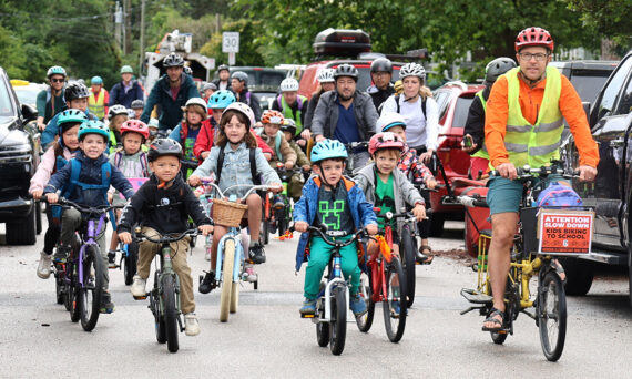 A large group of school-aged children riding bicycles together down a street in a Bike Bus, accompanied by a few adults who are also on bicycles.