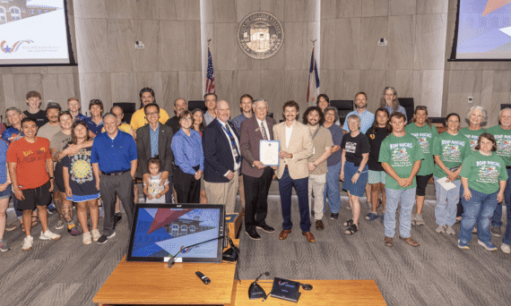 A large, diverse group of dozens of cyclists, including children in team jerseys, students, and adults in casual and riding gear, smiling for a crowded group photo inside a wood-paneled city council chamber.